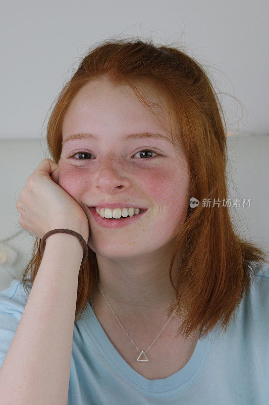 这是一个14 / 15岁的红发少女，皮肤苍白，脸上有雀斑，脸颊通红，坐在卧室里，看起来很开心，手托着下巴微笑着